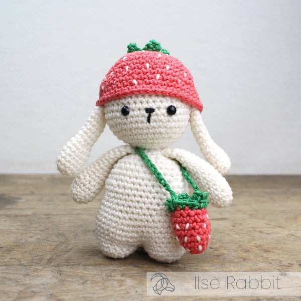 Crocheted rabbit toy with a pink hat and strawberry bag on a wooden surface